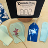 Cascade Press sunprint kit components laid out on a light wood surface, including a black press box, patterned blue and white tags, pale green blank tags, and a pressed flower sample.