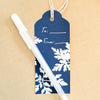 A navy blue gift tag with white leaf patterns and a white pen resting on top, set against a light beige textured background.