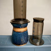 Ceramic vessel with AeroPress on top, on a blue surface with a neutral background