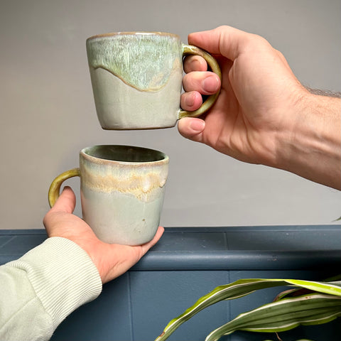 Two hands holding ceramic mugs with a dynamic green, yellow, and blue color combination.