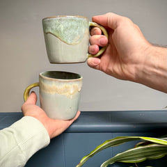Two hands holding ceramic mugs with a dynamic green, yellow, and blue color combination.