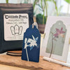 Cascade Press sunprint kit components displayed on a wooden surface, including a black pouch of fifteen gift tags, a navy blue tag with white floral artwork, and a sample printed card with a floral arch design.