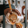 Hand pouring honey from a glass jar with a strainer into another container, blurred background