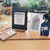 A DIY sunprint kit displayed on a wooden board on a beige table, with potted plants in the background. The kit includes a black pouch labeled Cascade Press Sunprint Kit, a sample sunprint card, a dark blue gift tag with a white floral print, and other small kit components.