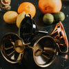 Three metal cocktail strainers on a dark surface with fruits in the background